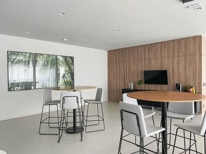 Een moderne eethoek met een ronde houten tafel en vier grijze stoelen. Een flatscreen televisie is aan de muur bevestigd en er is een groot raam met uitzicht op bomen.