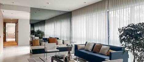 A modern living room with a blue sofa, white armchairs, and a coffee table. There is a large window with sheer curtains and a potted plant.