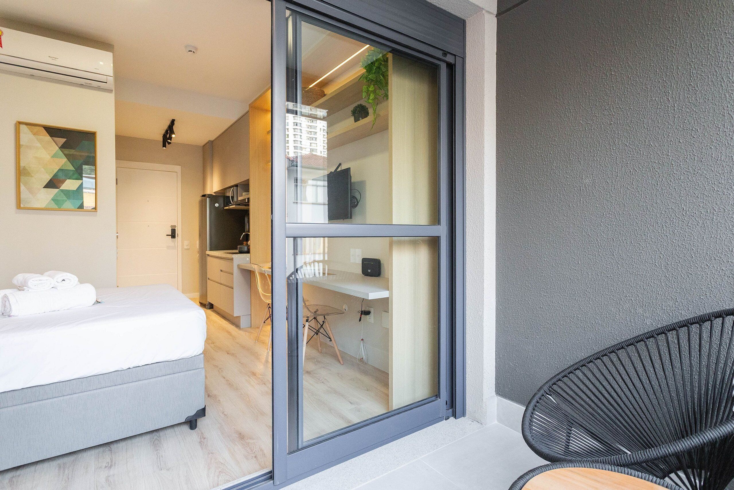 Una habitación moderna con una cama, una silla negra y un ventanal corredero que da a una zona de cocina.