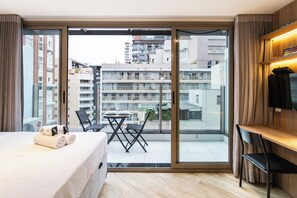 A modern hotel room with a large sliding glass door leading to a balcony, a bed with neatly folded towels, a small table with chairs, and a television on a wooden stand.