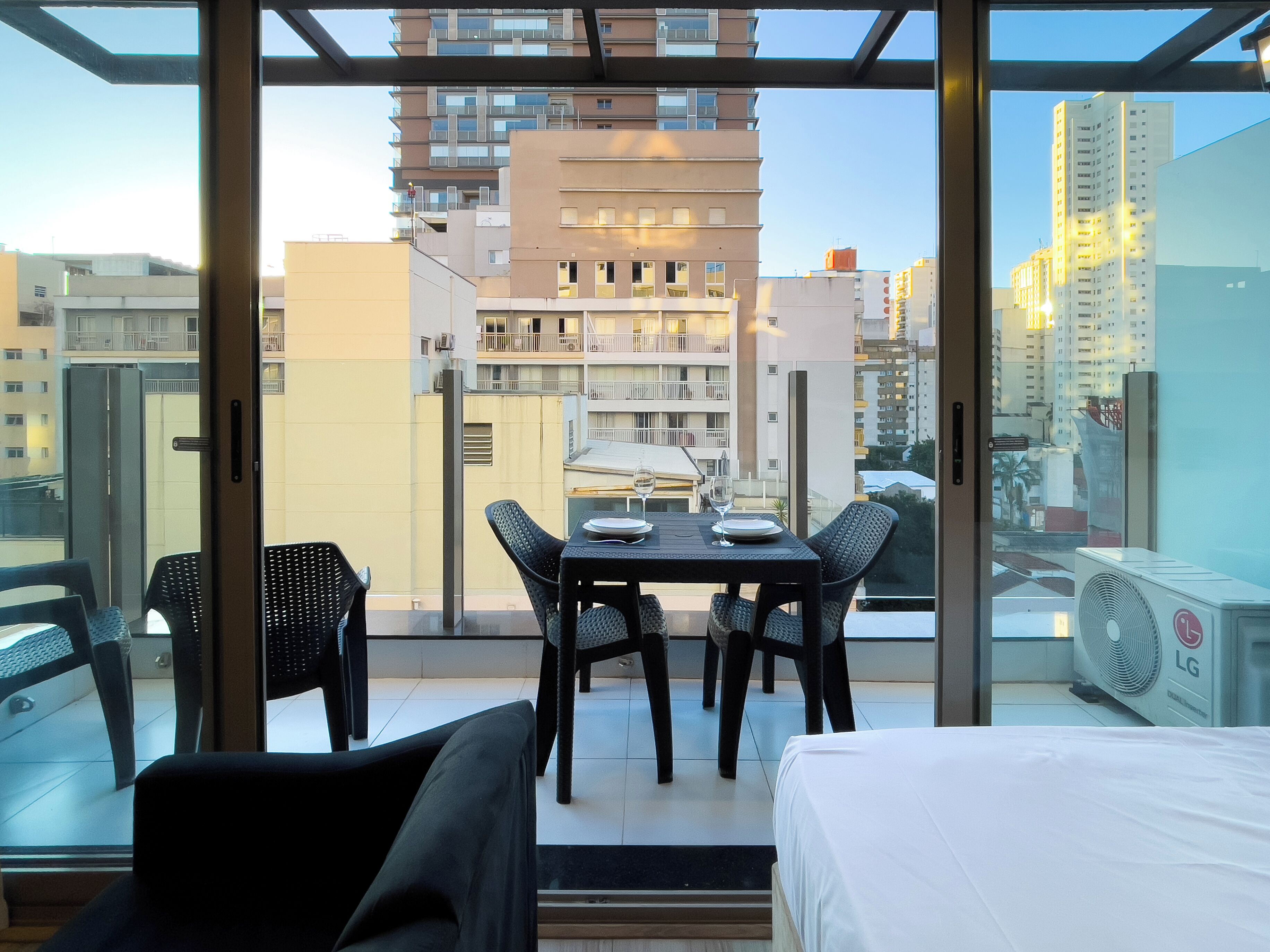 Varanda com mesa posta para dois, uma cama e vista dos edifícios ao redor.
