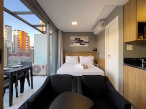 A modern hotel room with a bed, a dining area, and a view of the cityscape.