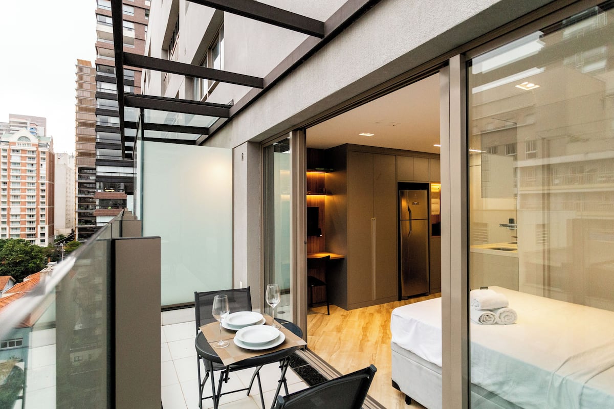 A modern hotel room with a balcony, a dining area with a table and chairs, and a bed with a neatly made white bedspread and rolled towels.
