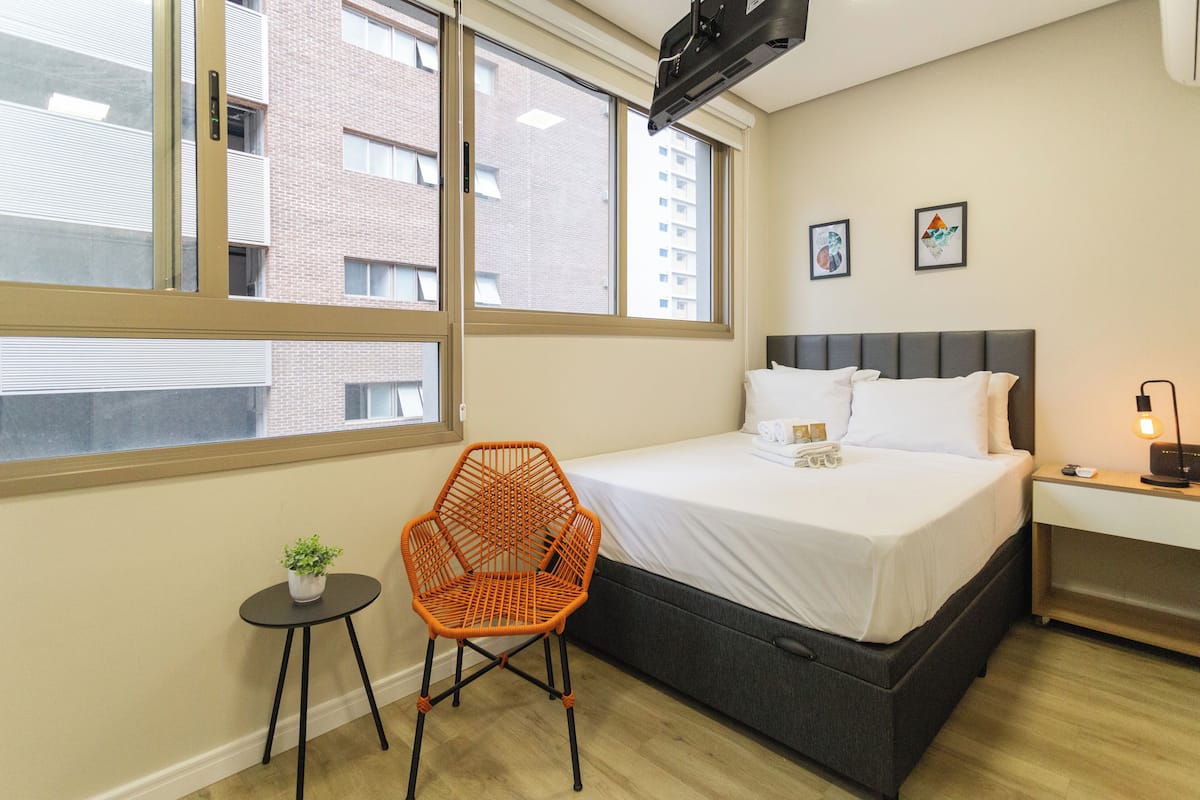 A bedroom with a bed, a small table with a plant, a chair, and a nightstand with a lamp.