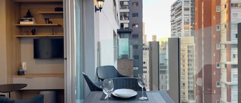 A balcony with a dining table set for two, a TV mounted on the wall, and a cityscape view.