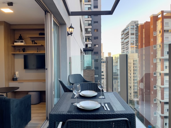 A balcony with a dining table set for two, a TV mounted on the wall, and a cityscape view.