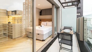 Moderni hotellihuone, jossa on sänky, pieni ruokailutila pöytineen ja tuoleineen, minikeittiö ja suuri ikkuna, josta avautuu näkymä kaupunkiin.