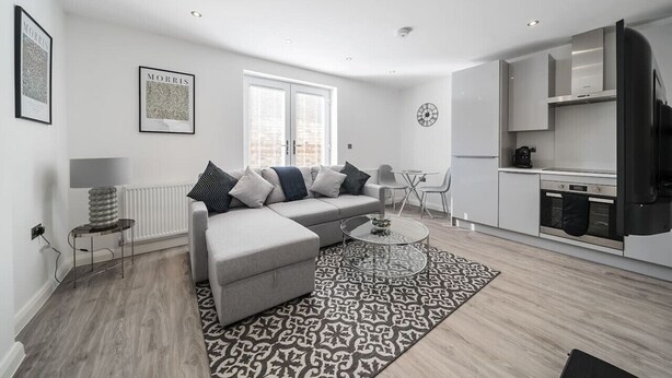 A modern living room with a grey sofa, patterned rug, and a kitchen area with white cabinets and a black refrigerator.