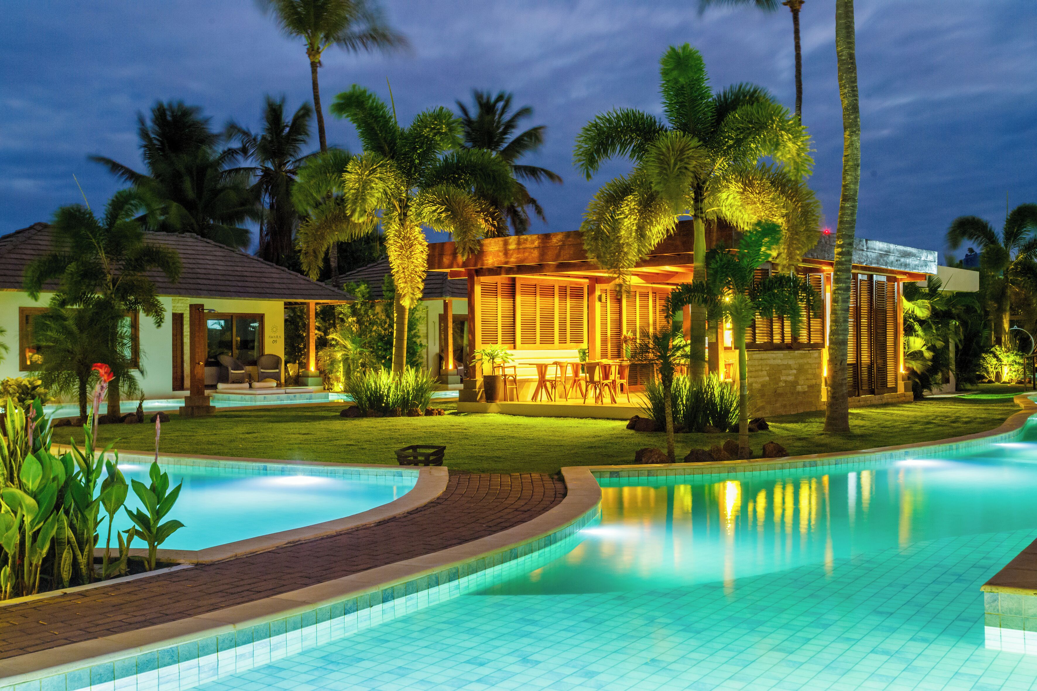 A swimming pool with a curved edge, surrounded by palm trees and a building with a patio area.