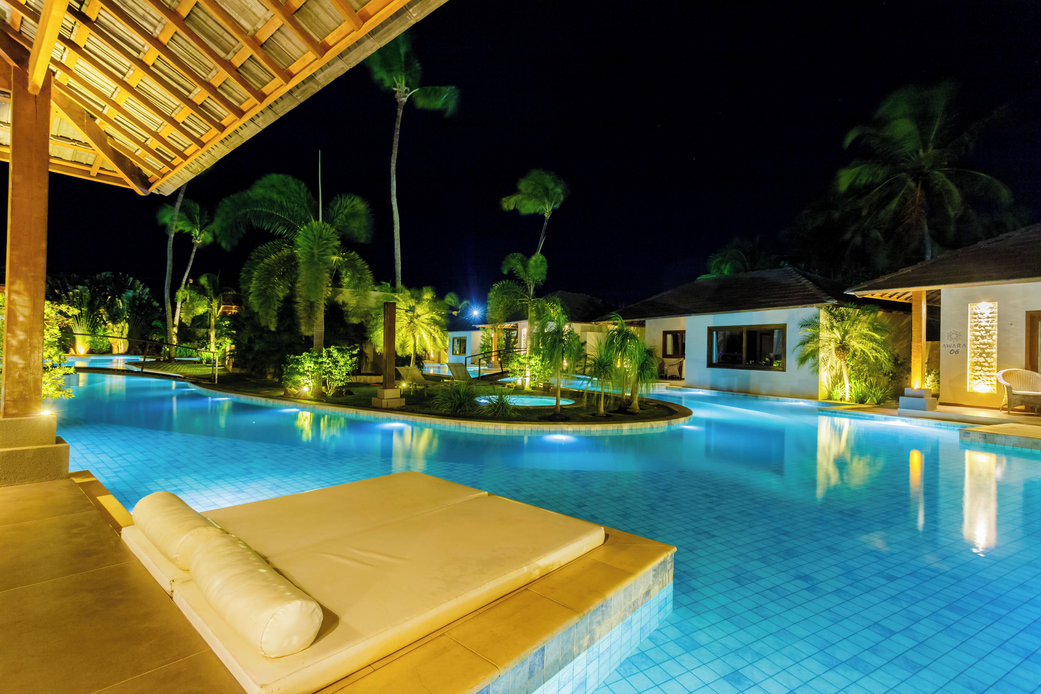 Un espace piscine nocturne avec des chaises longues, des palmiers et un coin salon abrité.