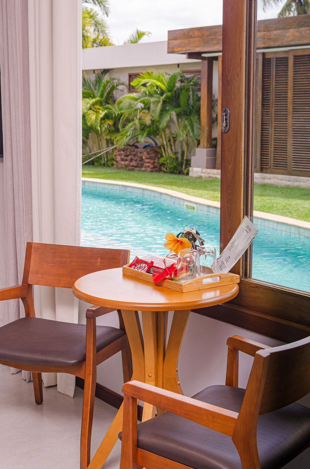A wooden table with a tray of snacks and a menu, two wooden chairs, and a view of a swimming pool with palm trees.