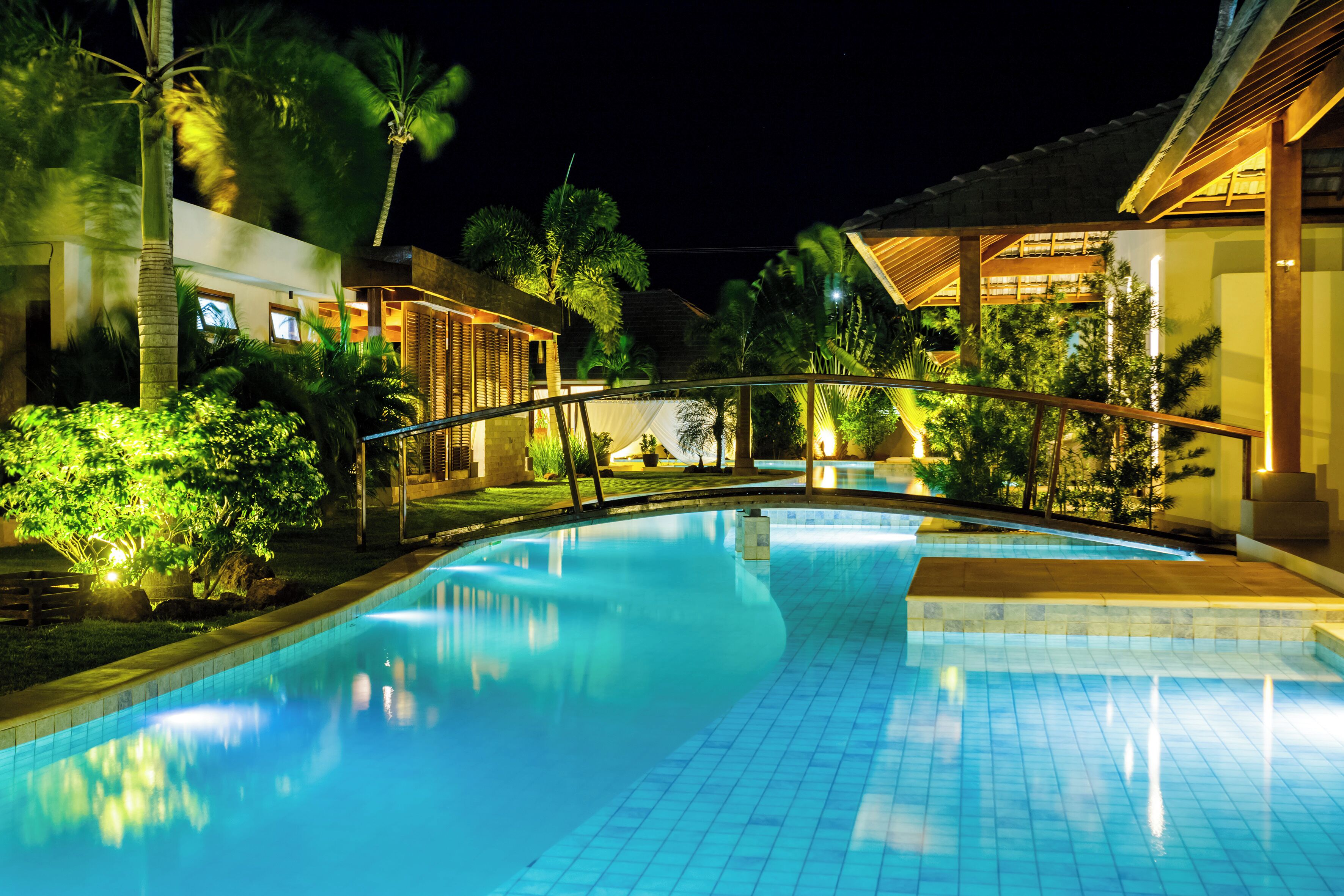 A nighttime view of a swimming pool with a bridge, surrounded by palm trees and buildings.