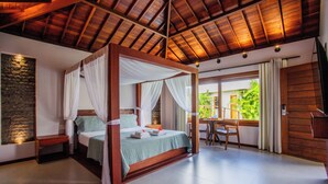 A four-poster bed with white drapes, a stone accent wall, a wooden desk, and a large window with a view of greenery. - Awara Pousada Boutique (São Miguel do Gostoso)