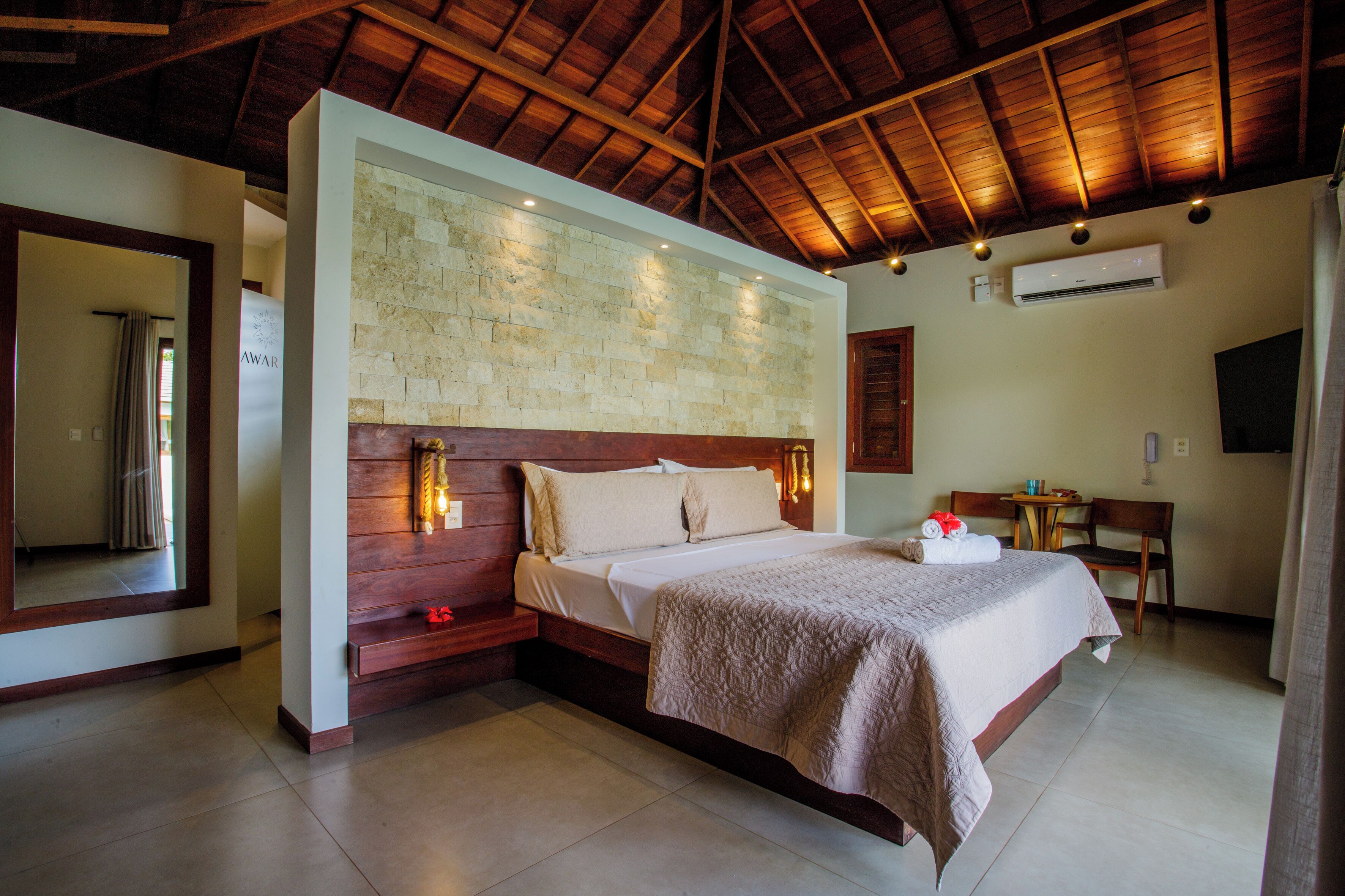 Un dormitorio espacioso con una cama grande, cabecera de madera y pared de piedra. En la esquina, hay una mesita y una silla, y un equipo de aire acondicionado en la pared.
