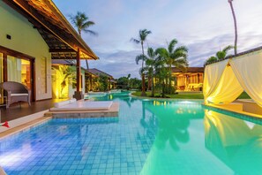 Una piscina con suelo de baldosas, un área de estar techada y palmeras al fondo.