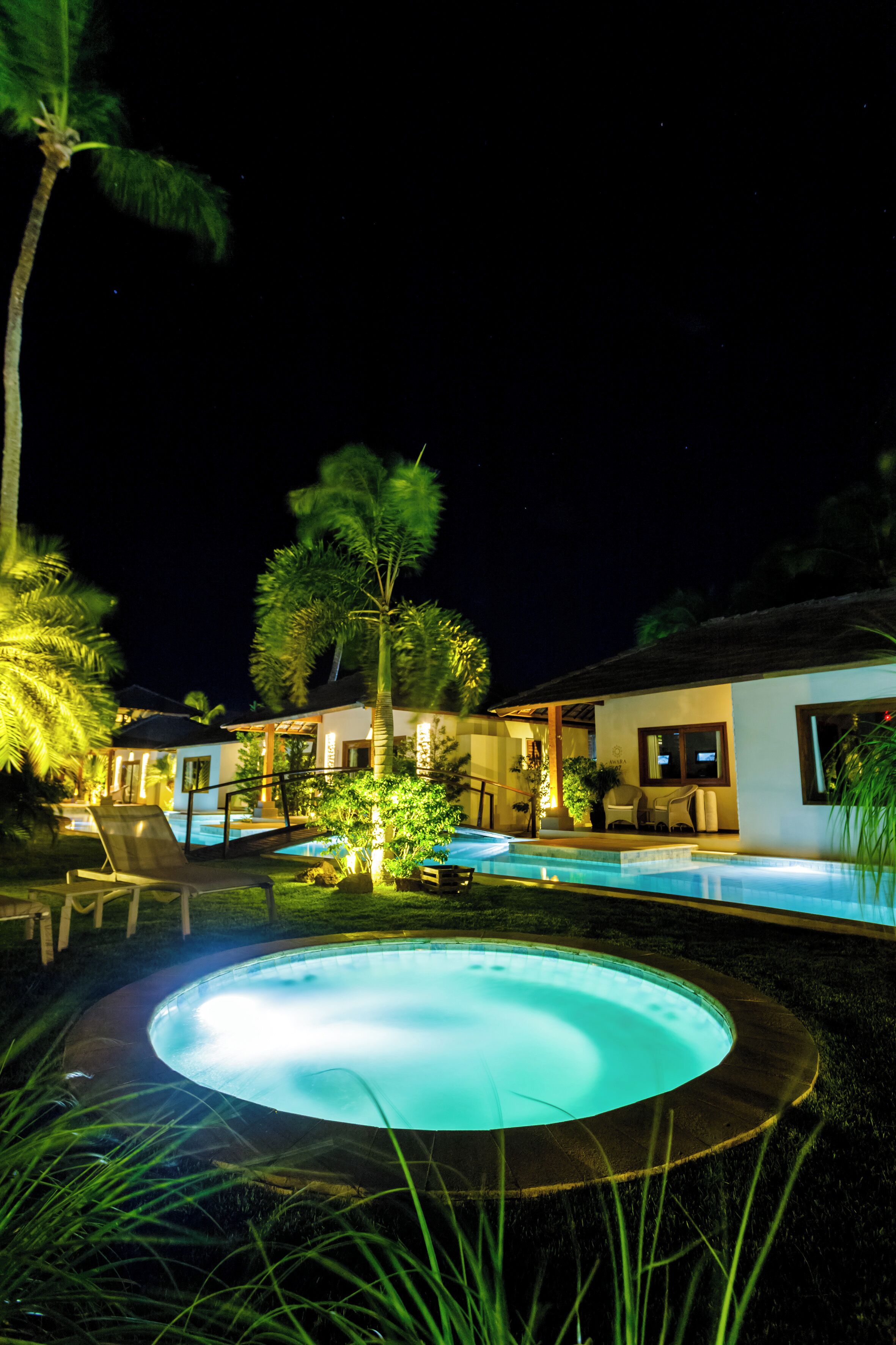 A nighttime view of a resort with a swimming pool, palm trees, and a building with a thatched roof.