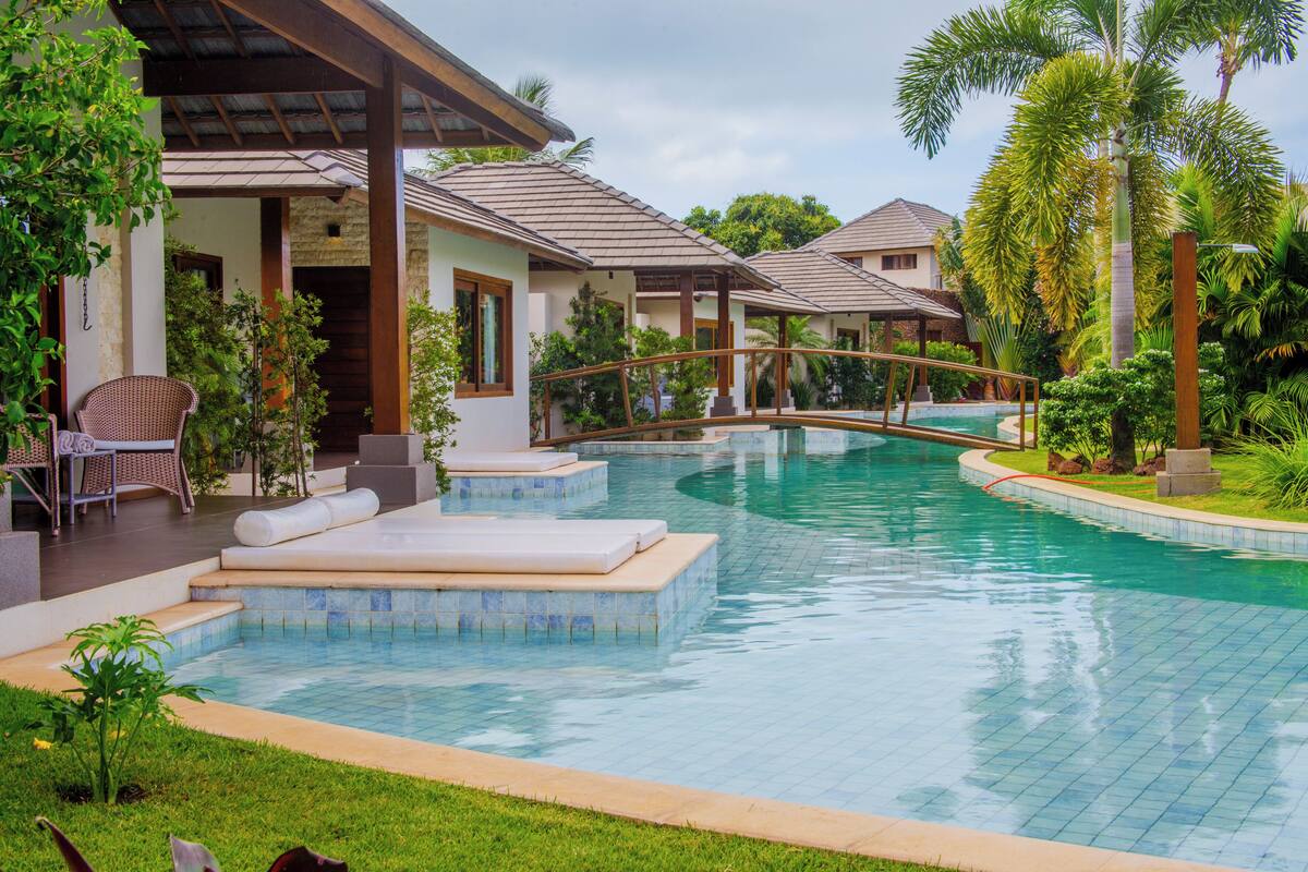 Una piscina con un puente, zona de descanso y jardines tropicales.