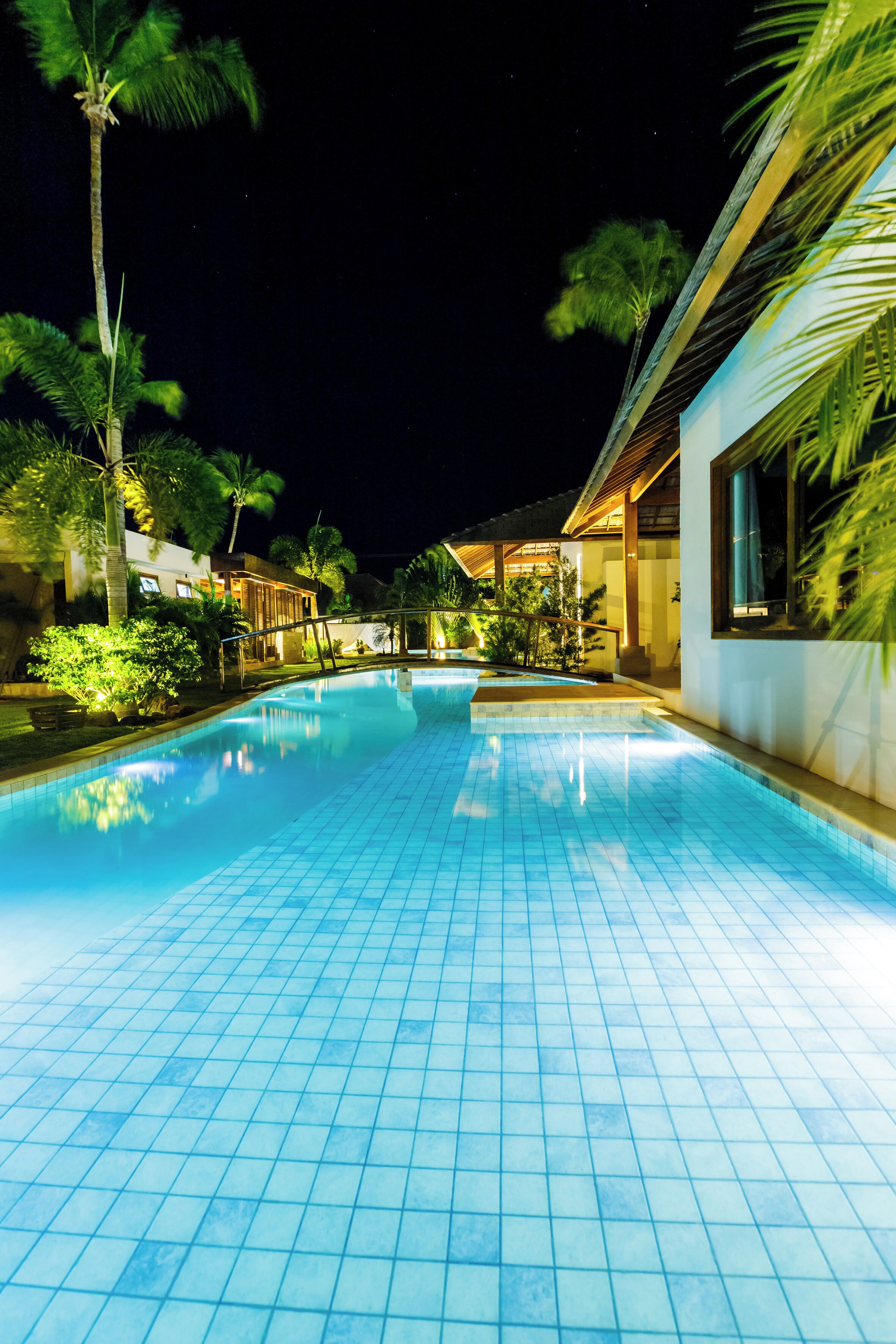 A nighttime swimming pool with tiled flooring, surrounded by palm trees and a building with large windows.