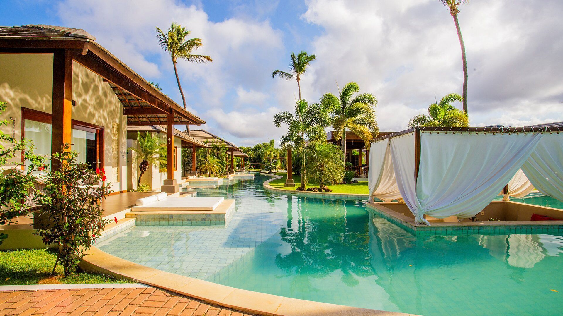 A resort pool area with a clear pool, palm trees, and a covered seating area.