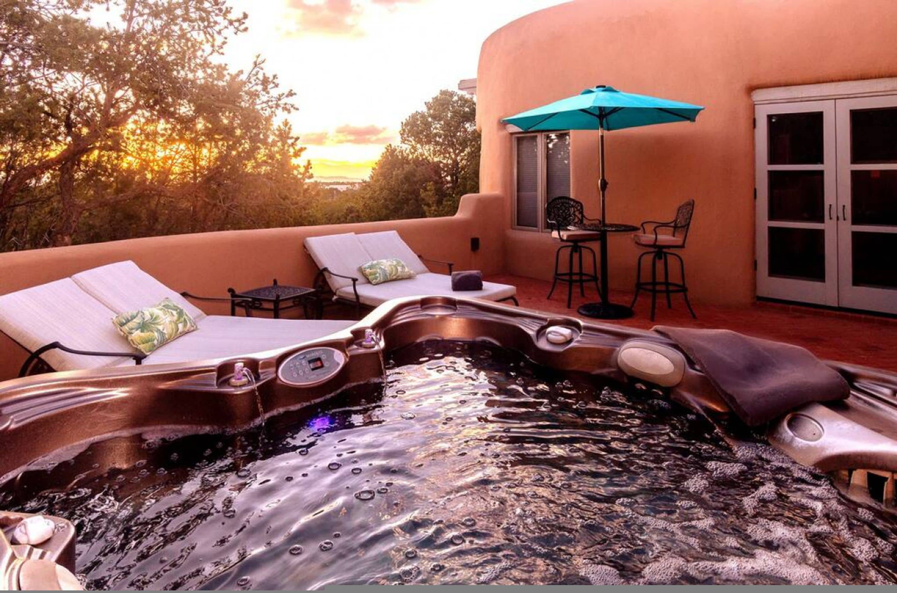 A hot tub with lounge chairs and a table, surrounded by a peach-colored building with a glass door.