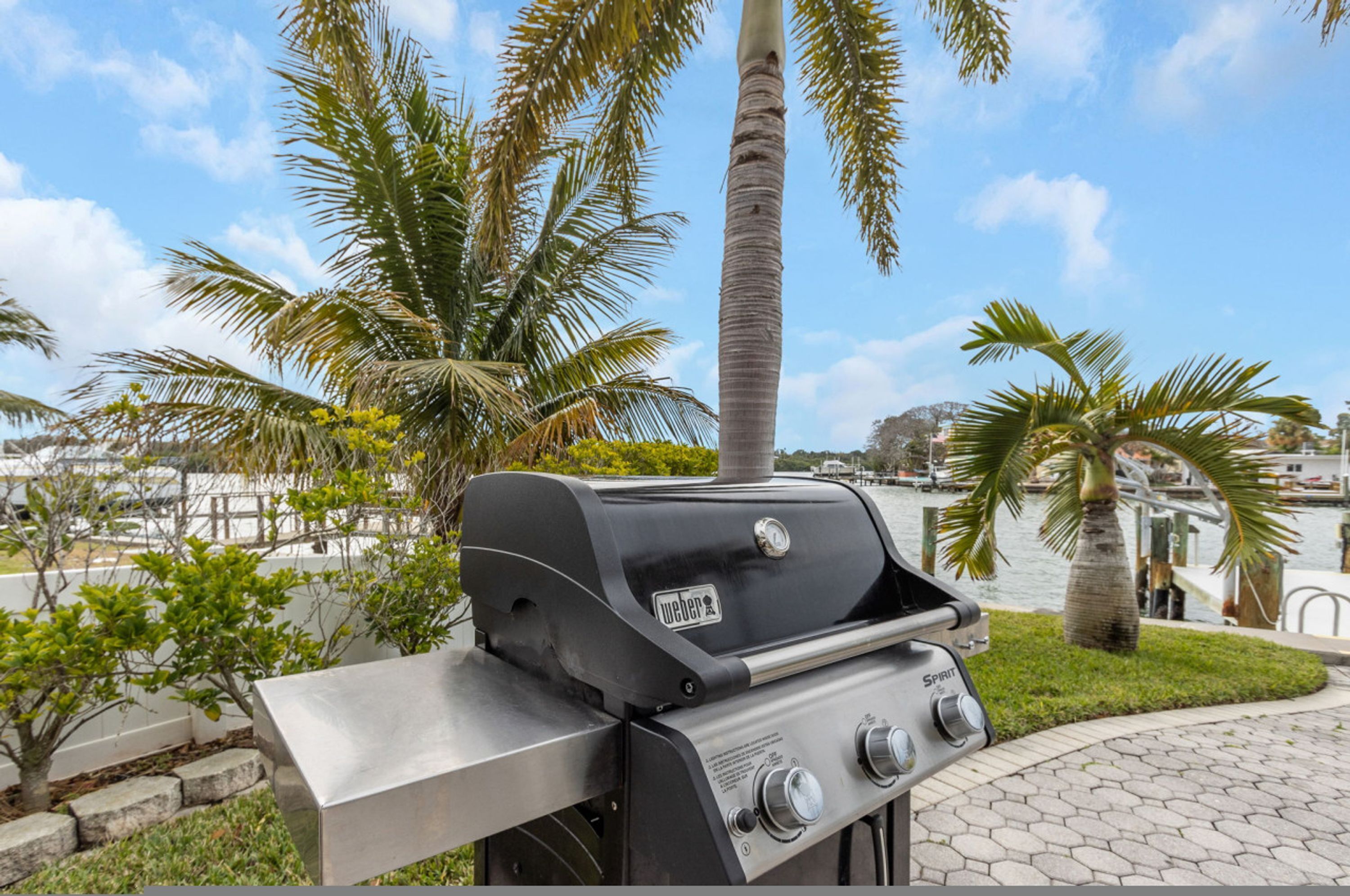 Ein Weber Spirit Grill mit Edelstahl-Oberfläche, in einem begrünten Außenbereich mit Palmen und Blick auf eine Marina.