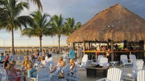 Ein Außenbereich mit weißen Stühlen und einem strohgedeckten Gebäude, vermutlich eine Bar oder Lounge, mit Palmen im Hintergrund.