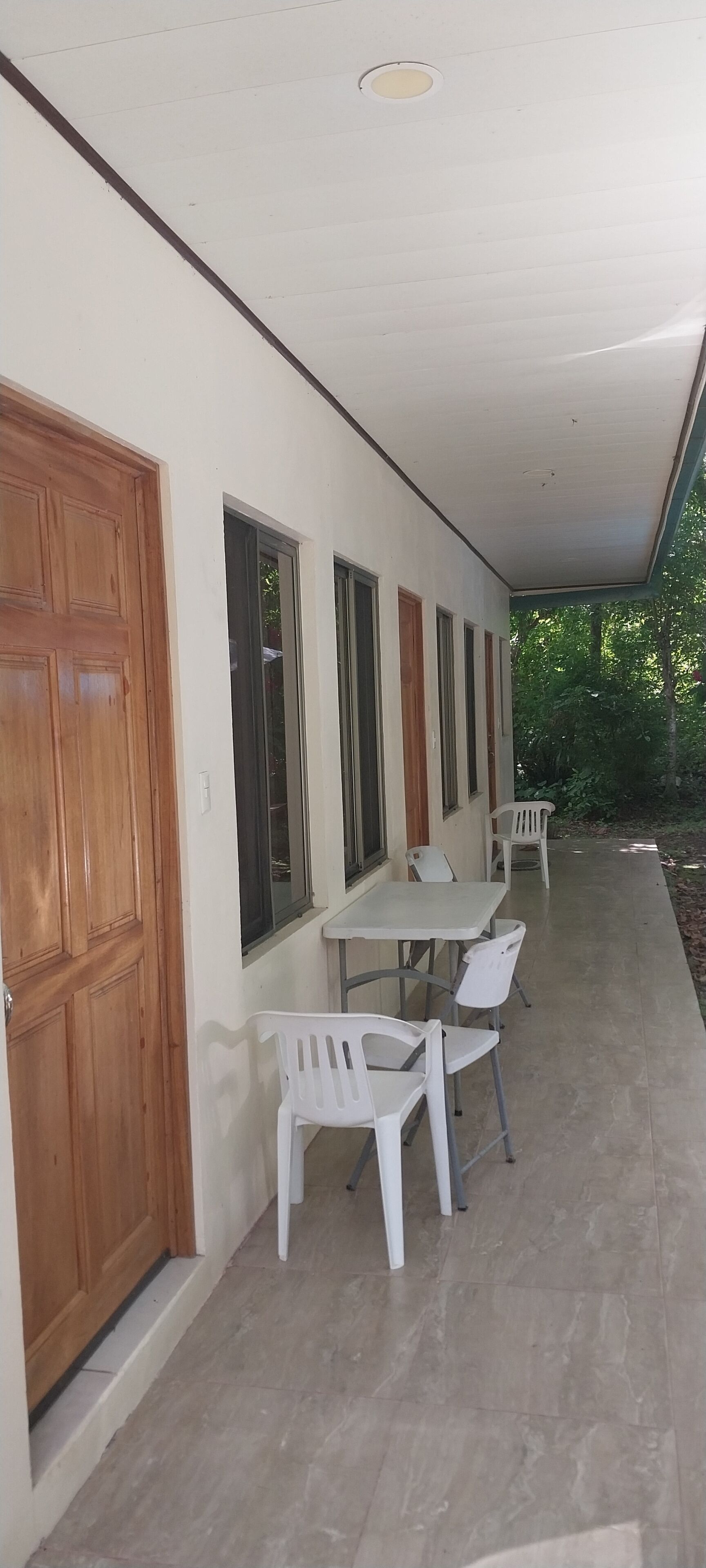 Un espacio al aire libre techado con una mesa, sillas, una puerta de madera y ventanas a lo largo de la pared.