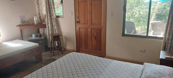 A bedroom with a bed, a wooden door, a window with a view of outdoor chairs, and a small table with a kettle and cups.