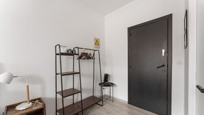 A room with a wooden nightstand, a desk lamp, a metal shelving unit, a black door, and a small table with a chair.