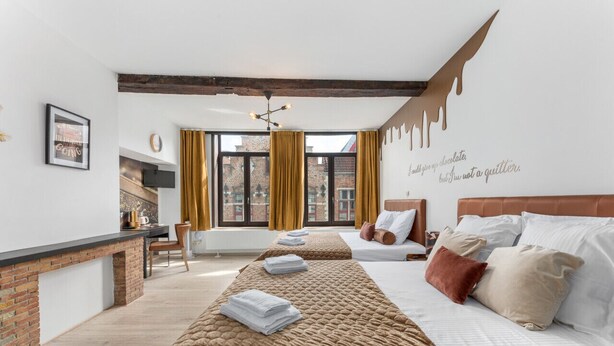 A hotel room with a long bed, a smaller bed, a fireplace, and a large window with curtains.