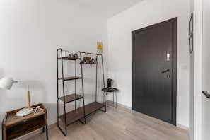 Una habitación con mesita de noche de madera, lámpara de escritorio, estantería metálica, puerta negra y una pequeña mesa con silla.