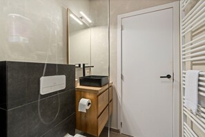 Un bany amb una paret revestida de rajoles negres, un tocador de fusta, un lavabo negre, una porta blanca i un portaTovallons.