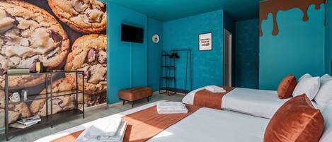Habitación de hotel con dos camas, un mural en la pared de galletas, un televisor de pantalla plana, una escalera y un letrero que dice 'Galletas de chocolate'.