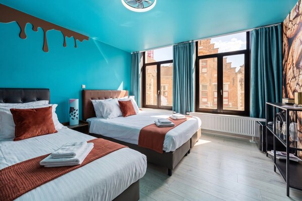 Une chambre d’hôtel avec deux lits, une grande fenêtre avec des rideaux, un radiateur et une vue sur les bâtiments environnants.
