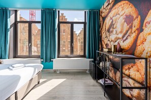 Een hotelkamer met een bed, een radiator, een raam met gordijnen en een muur met een afbeelding van een koekje.