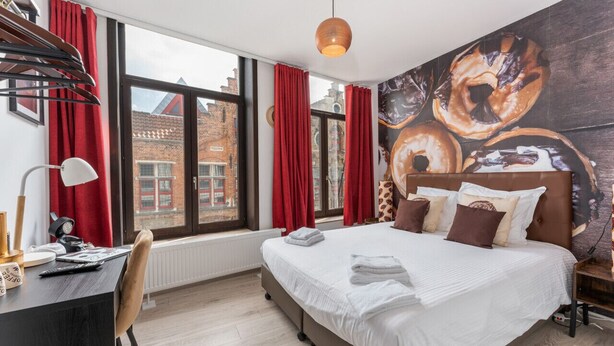 A modern hotel room with a large bed, a desk with a lamp, and a view of the city through large windows. - Homer Simpson's Donut Room - Steps from the Belfry (Brugge)