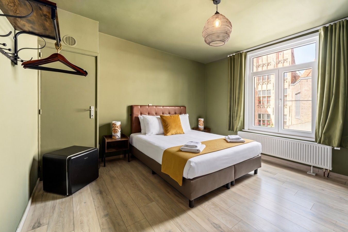 Une chambre d’hôtel avec un lit, des tables de chevet, un luminaire suspendu, un radiateur et une fenêtre avec des rideaux.