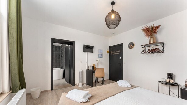Quarto de hotel com cama, escrivaninha, cadeira, pequena mesa com cafeteira, prateleira com objetos decorativos e chuveiro.