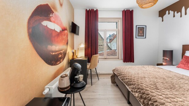 Une chambre d’hôtel moderne avec une grande fresque murale, un lit recouvert d’une couverture à motifs, un bureau avec une machine à café, une chaise et un téléviseur.