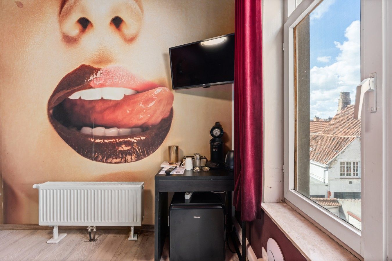 A room with a large wall mural, a radiator, a desk with a TV, and a window with a view of buildings.