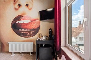 Ett rum med en stor väggmålning, ett element, ett skrivbord med en tv och ett fönster med utsikt över byggnader.