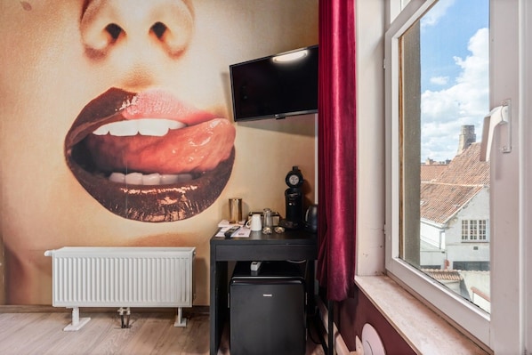 Una habitación con un mural grande en la pared, un radiador, un escritorio con televisor y una ventana con vistas a edificios.