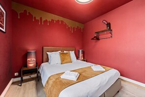 Habitación de hotel con pared roja, cama con ropa de cama blanca y marrón, dos lámparas de noche y un estante fijado a la pared.