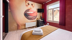 Een hotelkamer met een grote muurschildering, een bed met een witte deken, een radiator, een bureau met een stoel en een raam met gordijnen.
