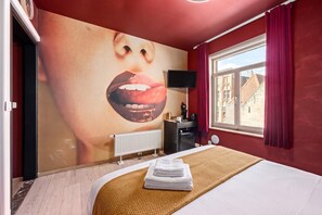 A hotel room with a large wall mural, a bed with a white comforter, a radiator, a desk with a chair, and a window with curtains. - The Hot Chocolate Room by Charlie`s Choco Factory (Brugge)