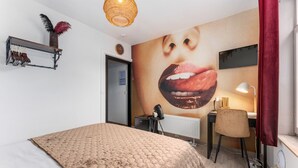 Une chambre avec un grand lit, un bureau avec une chaise, une étagère à chaussures, un téléviseur fixé au mur et un rideau rouge.