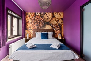 Habitación de hotel con una cama grande, paredes moradas, una ventana con cortinas y un mural de galletas.