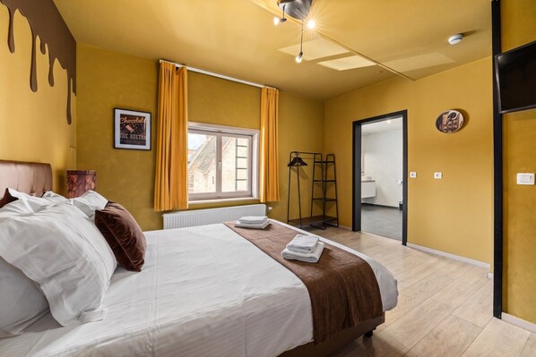 A hotel room with a bed, a window with curtains, a radiator, a mirror, and a door leading to another room.