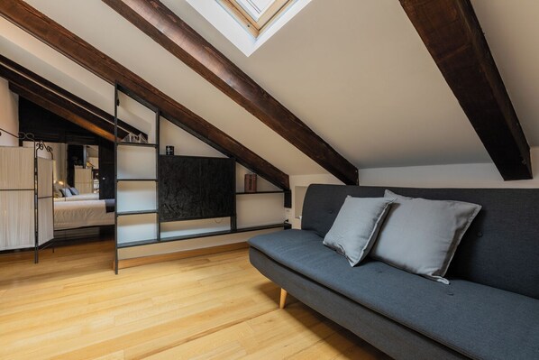 Ein Schlafzimmer im Loft-Stil mit schrägem Dach, Holzbalken, einem grauen Sofa und einer integrierten Regallösung.