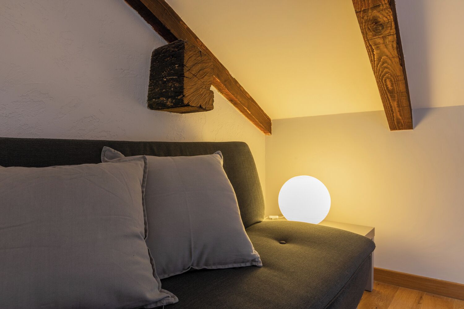 Um quarto aconchegante com um sofá cinza escuro, duas almofadas cinzas e uma luminária redonda fixada na parede.